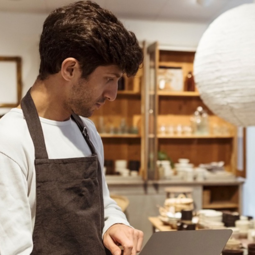 shop worker wearing an apron and reading a tablet with customers in the background