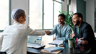 Image of two people sitting across a desk from a woman. The two woman are shaking hands.