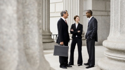 三位身着黑色西装的高管站在大厅里聊天，周围是巨大的柱子。