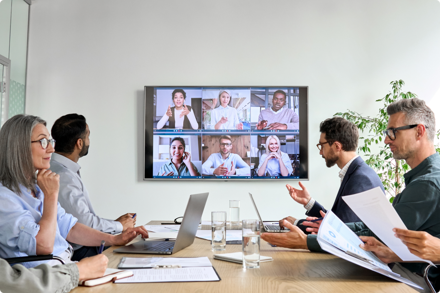 Group of people in a meeting at a table looking at a TV with remote video conferencing.