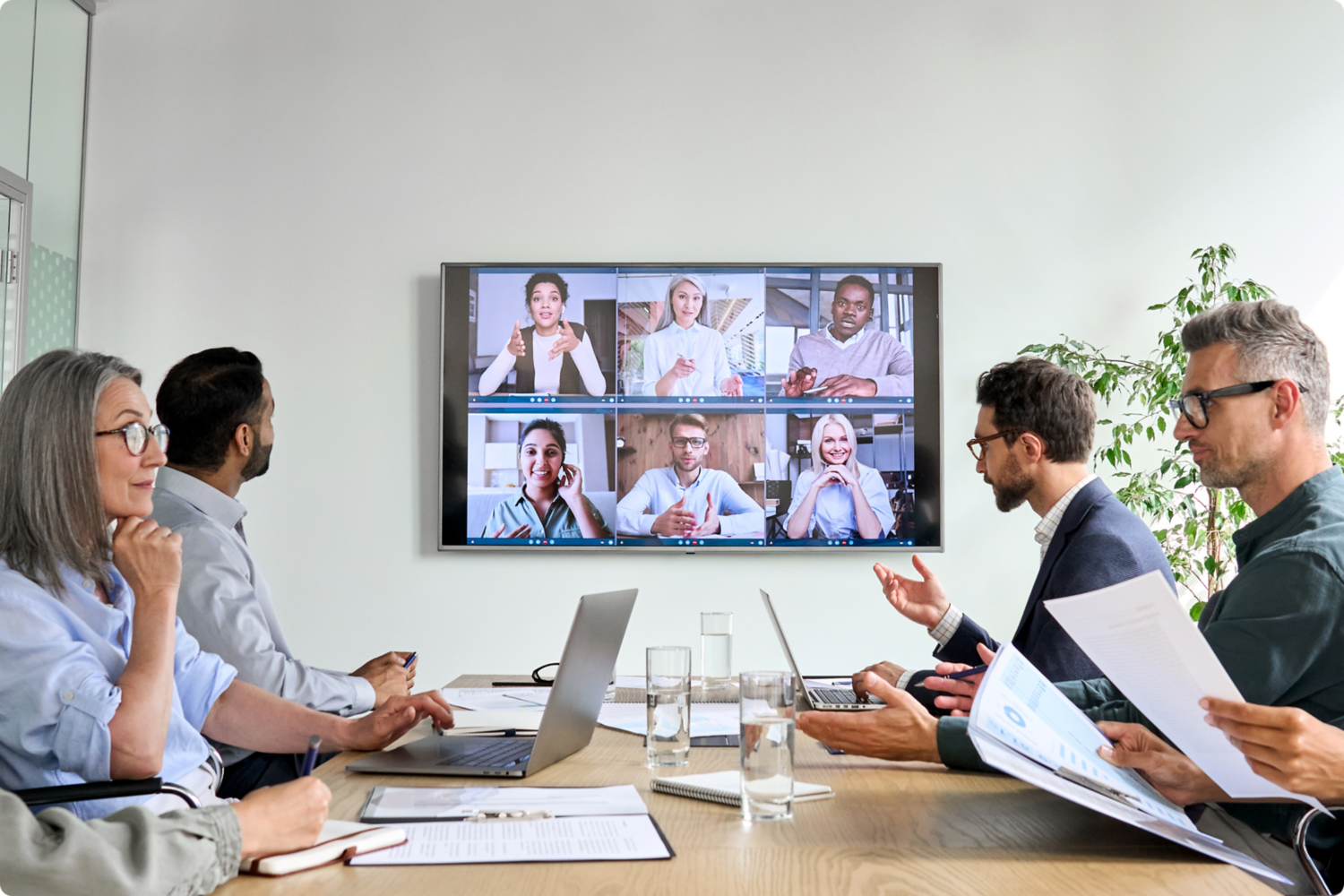 Gruppo di persone impegnate in un meeting a un tavolo che guardano una TV con videoconferenza in remoto. 