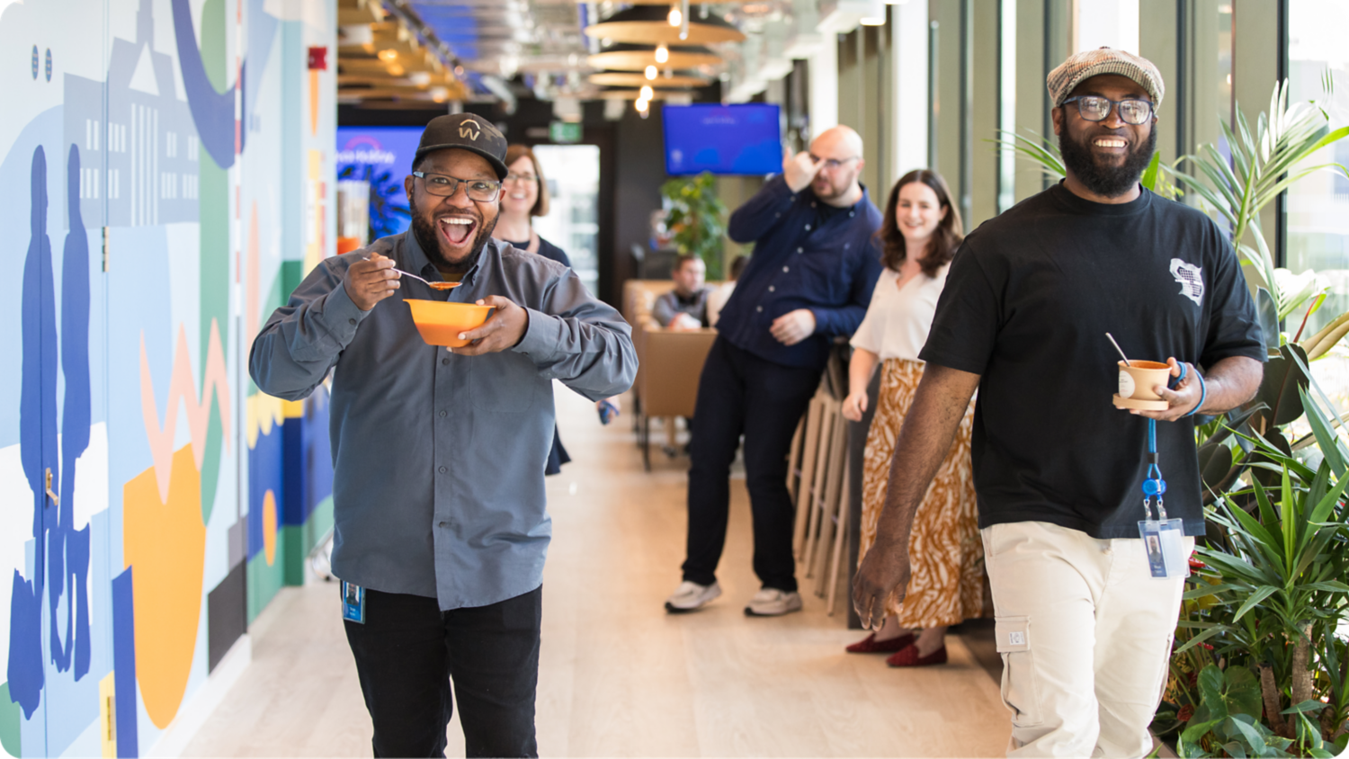 Two employees walking down a hallway, holding their lunches and laughing.