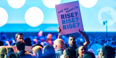 Workday employee holding a "Are you ready to rise?" sign in a filled auditorium at Workday Rising.