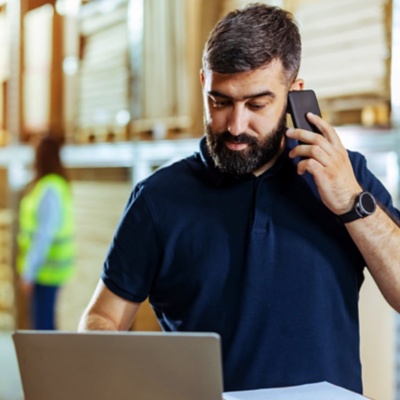 Employee in warehouse working on a laptop and holding a mobile phone